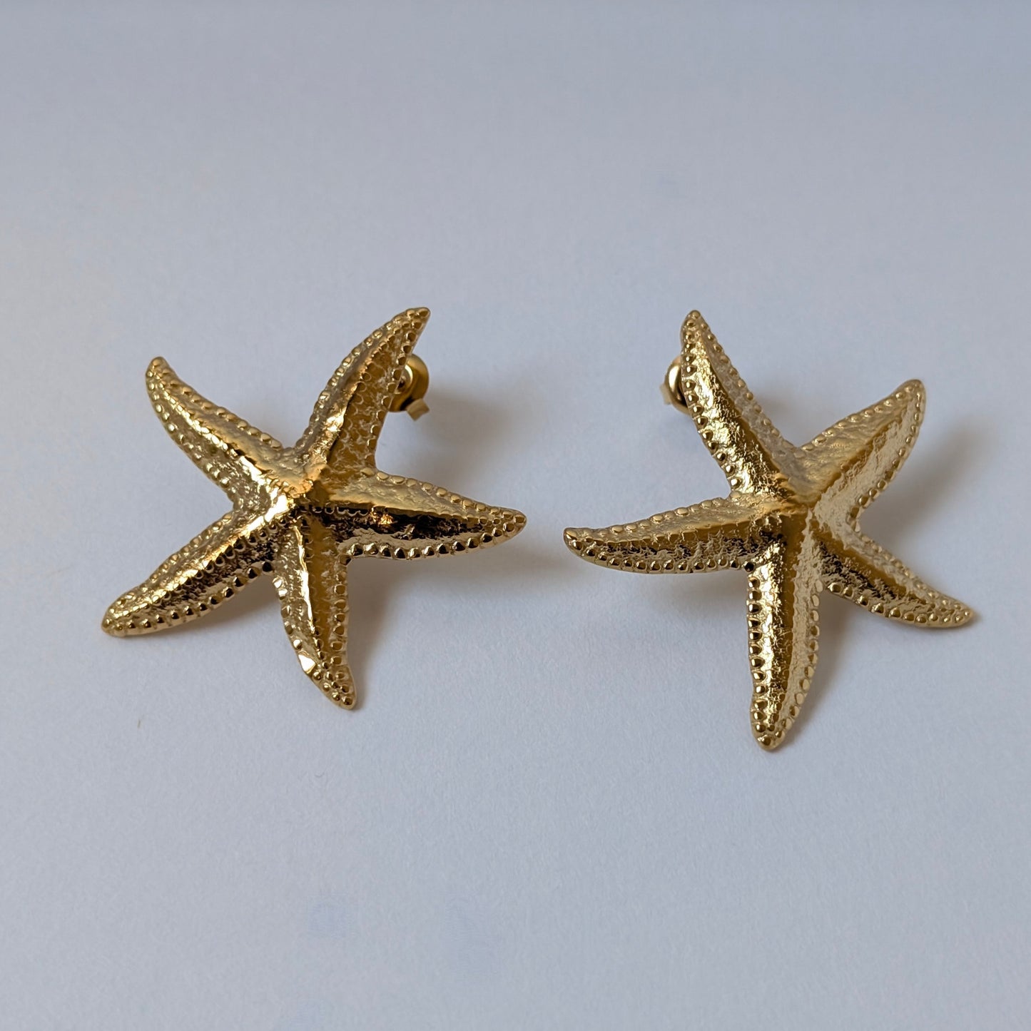 Alternate flat‑lay of gold starfish stud earrings angled to show texture, beside a seashell prop on white background.