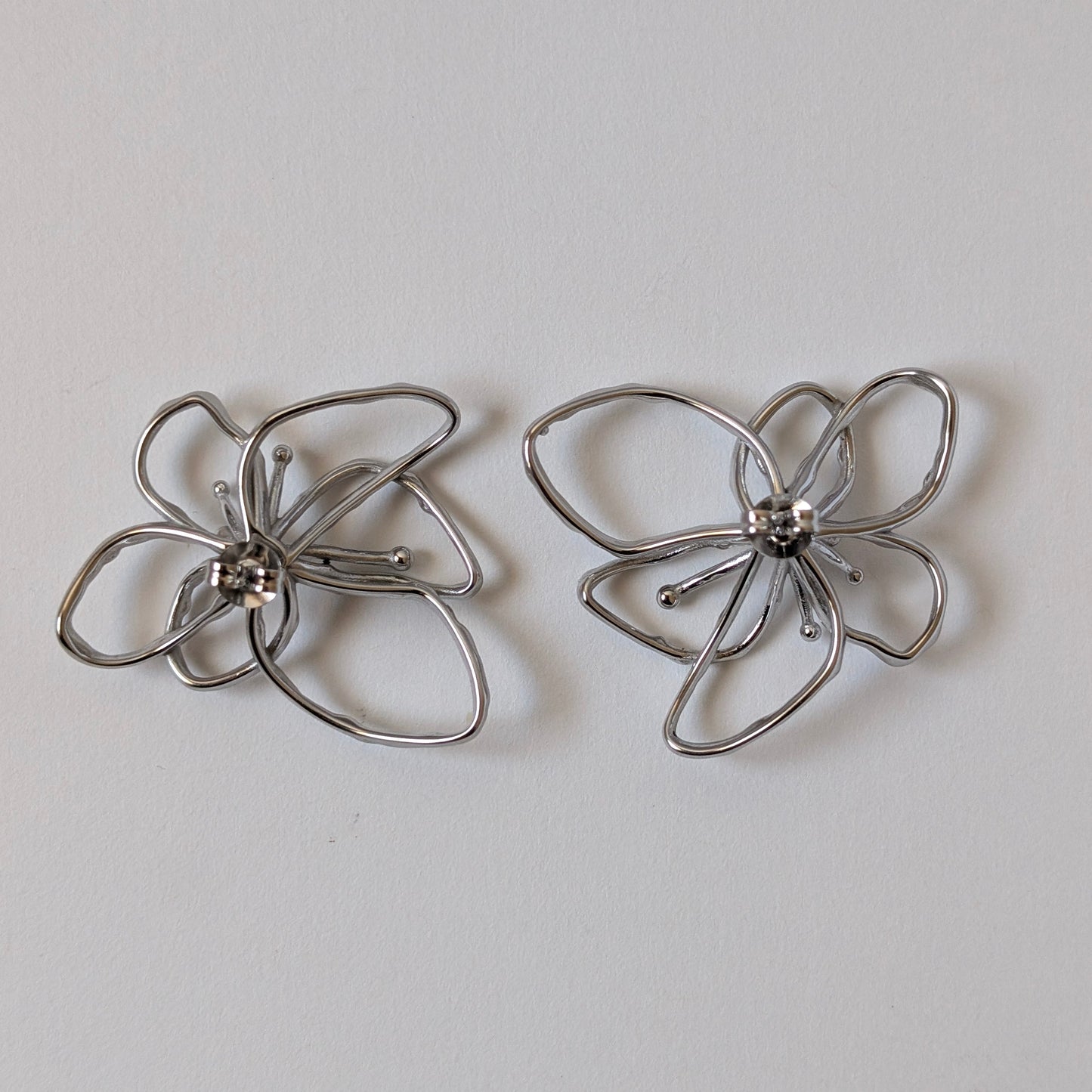 Flat‑lay of silver orchid statement earrings front side up, posts facing down on white backdrop—316 L steel petals.